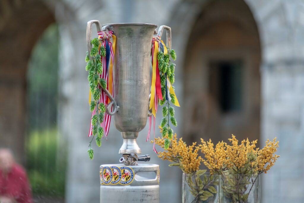 A silver trophy cup adorned with hop vines and ribbons, resting on a metal keg base labeled with Maryland Brewers Association stickers.