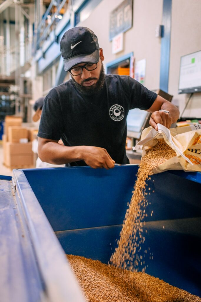 A man wearing a black Sankofa Beer Company t-shirt and cap pours a bag of grain into a large blue bin.