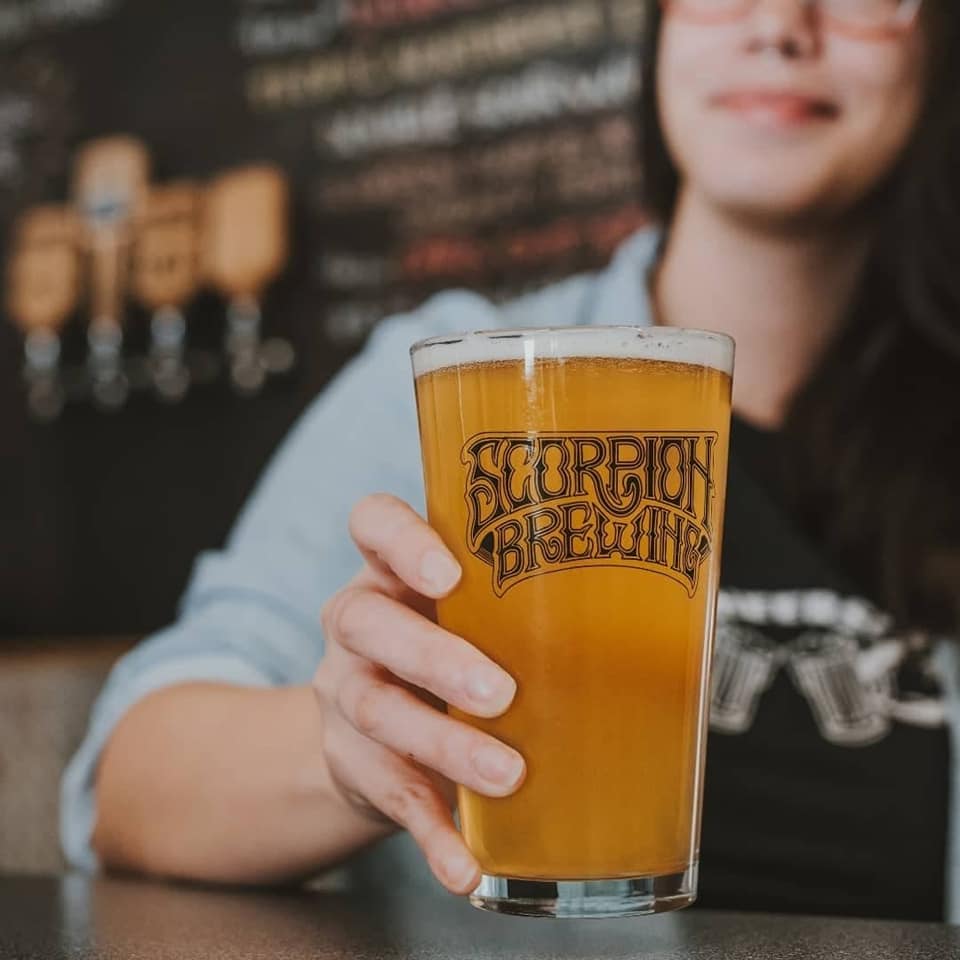 A person holds a glass of beer labeled with the Scorpion Brewing logo.