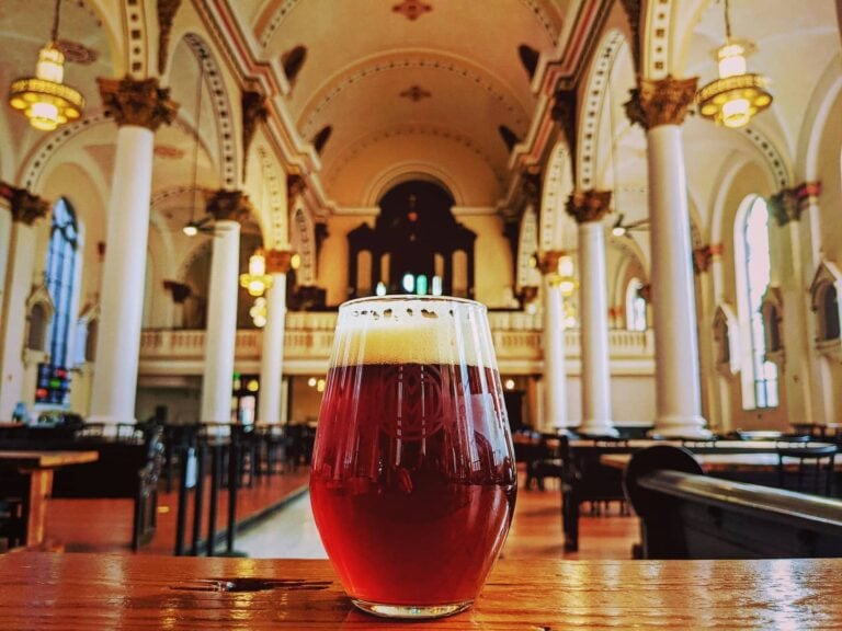A glass of amber beer sits on a wooden bar inside a spacious hall with white pillars and high arched ceilings.