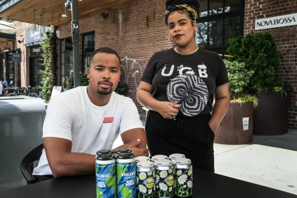 A man and a woman standing at an outdoor table with six cans of craft beer.