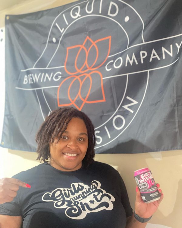 A smiling woman wearing a “Girls Running Sh*t” t-shirt holds a can of She’s Intense beer in front of a Liquid Brewing Company flag.
