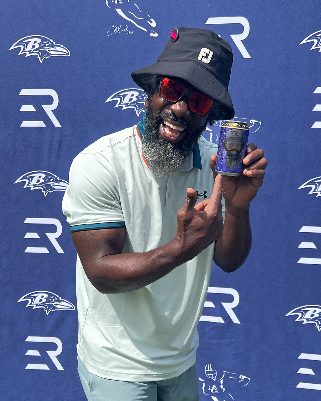 Ed Reed wearing a black bucket hat and sunglasses, smiling while pointing at a canned beverage featuring his own image.