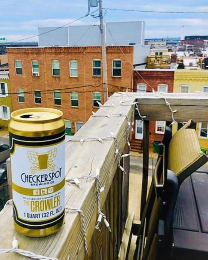 A Checkerspot Brewing Co. crowler can resting on a wooden rooftop railing, with urban brick buildings in the background.