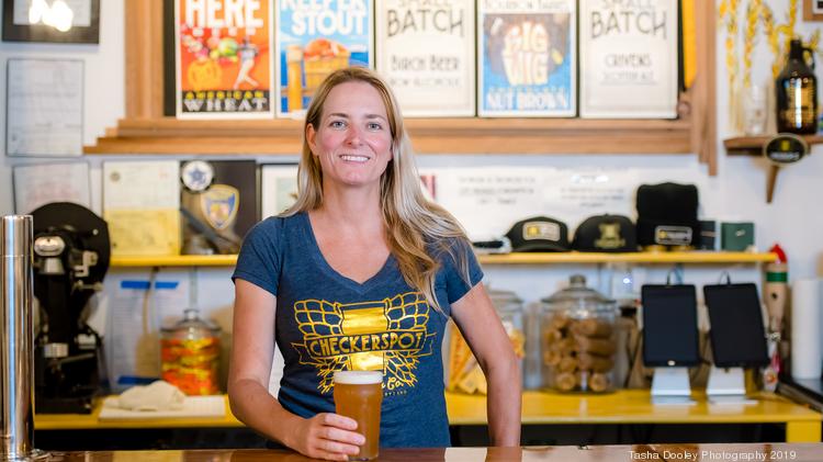 A smiling woman in a branded t-shirt holds a glass of beer at a bar with beer advertisements and signage in the background.