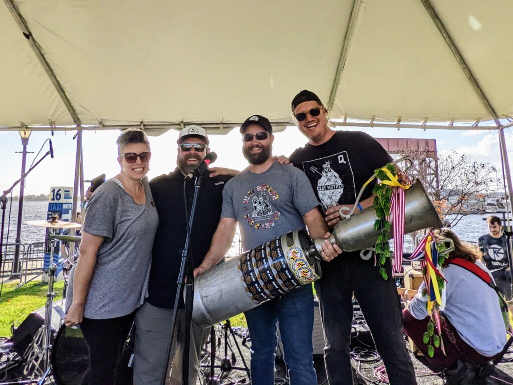Four people standing under a tent on a stage, with one holding a large trophy made from a keg decorated with ribbons.
