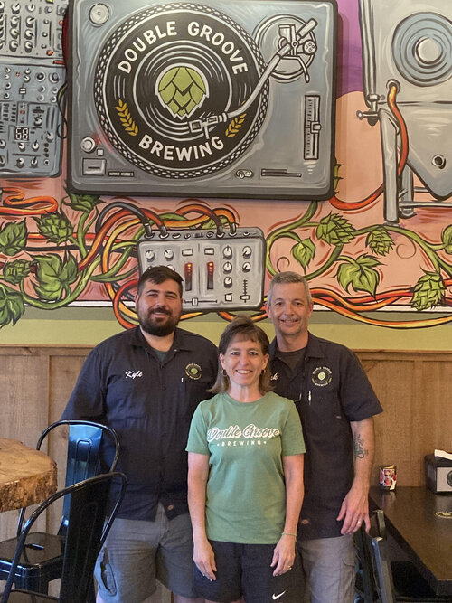 Three people standing in front of a mural depicting a record player and brewing equipment, with the Double Groove Brewing logo.