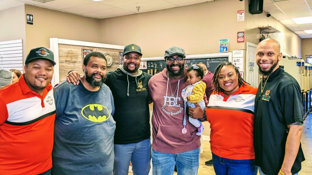 A diverse group of six adults stands together in a brewery, with one man in the center holding a baby.