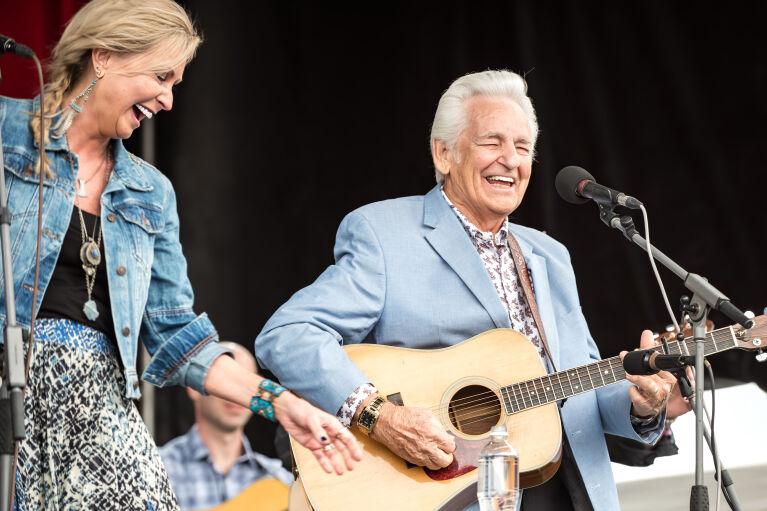 A woman laughing and an older man in a blue blazer smiling while playing an acoustic guitar on stage.
