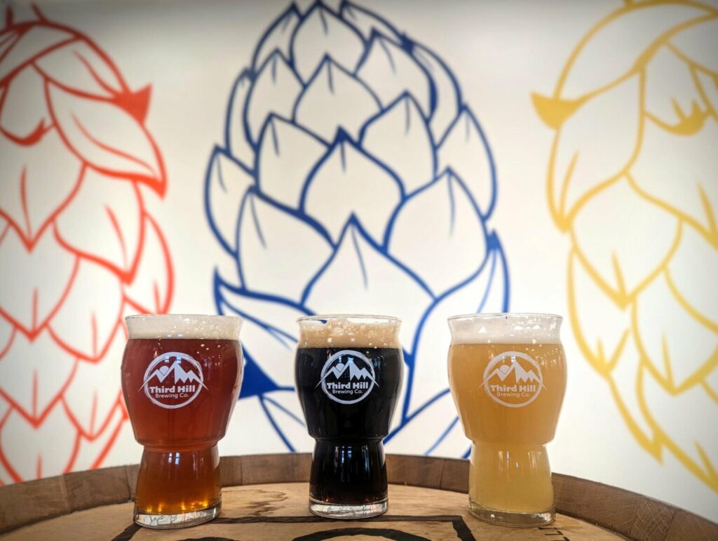 Three glasses of beer in different colors—red, dark brown, and golden—each featuring the Third Hill Brewing Co. logo, sitting on a wooden barrel in front of a wall mural of hop cones.