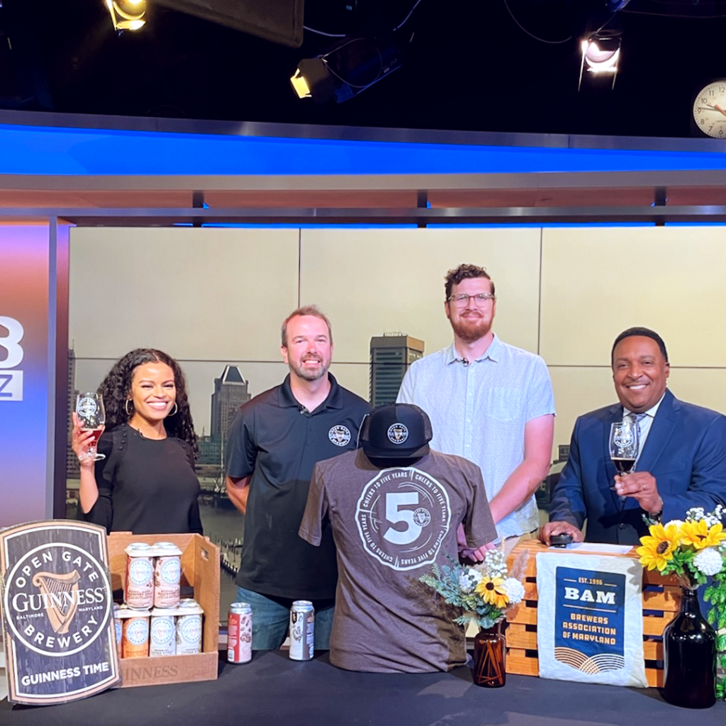 Four people stand behind a table featuring Guinness cans, a Brewers Association of Maryland banner, and a commemorative t-shirt marking five years.