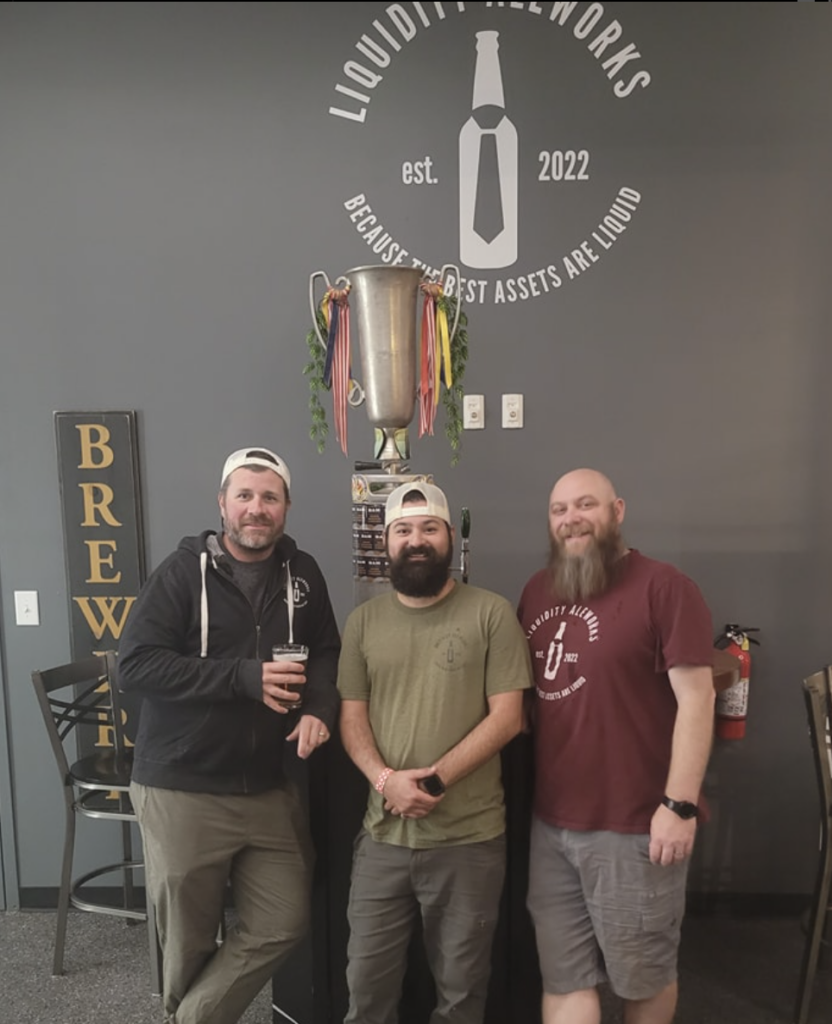 Three men stand in front of a Liquidity Aleworks logo and a large trophy.