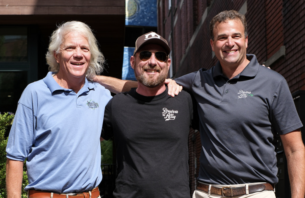 Three men with arms around each other’s shoulders, wearing shirts with the Brewers Alley logo.