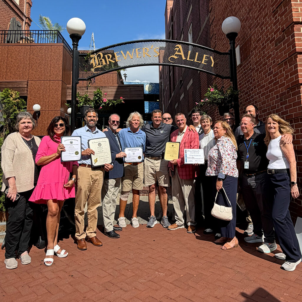 A group of people standing under a “Brewer’s Alley” archway, with several individuals holding framed certificates.
