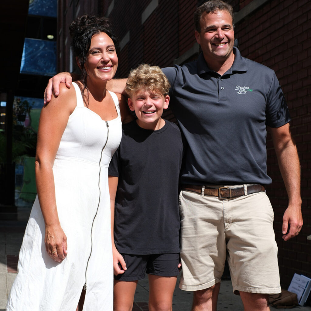 A smiling woman in a white dress, a young boy in a black t-shirt, and a smiling man in a grey polo shirt and khaki shorts stand together outdoors.