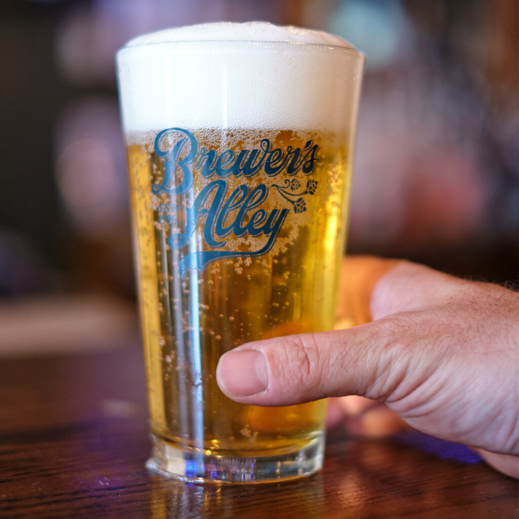 A hand holds a glass of beer with a thick white head, featuring the “Brewer’s Alley” logo printed on the side.