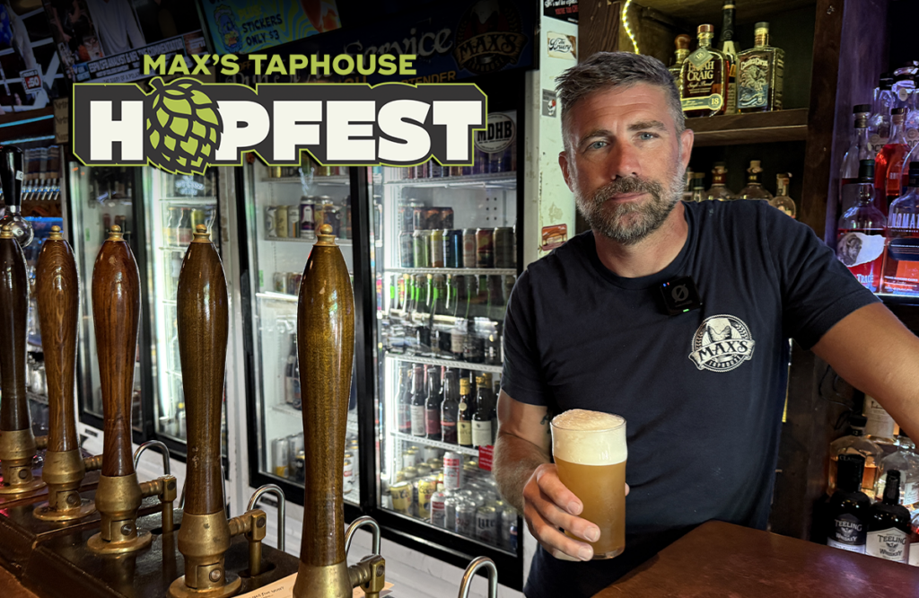 A man in a Max’s Taphouse t-shirt holds a glass of beer behind a bar featuring a Max’s Taphouse Hopfest sign.