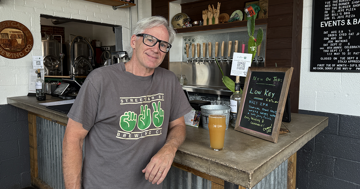 A smiling man wearing a Streetcar 82 Brewing Company t-shirt leans on a bar next to a glass of beer and a chalkboard sign listing a hazy IPA.