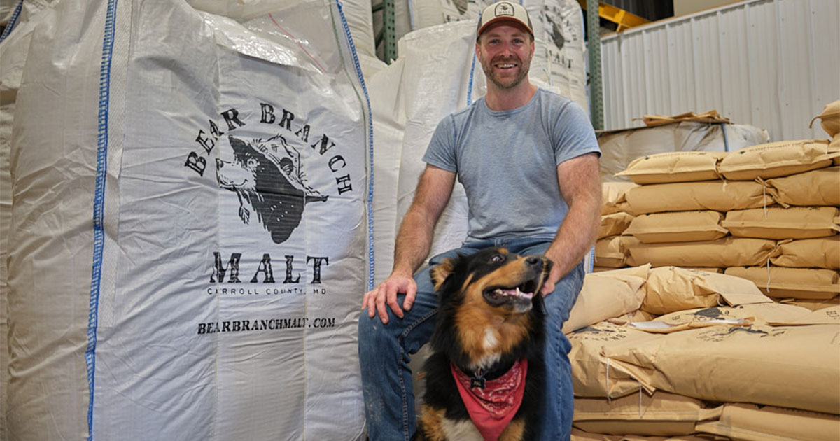 A man wearing a baseball cap and his dog sit in a warehouse next to a large white bag branded with the Bear Branch Malt logo and stacks of smaller grain bags.