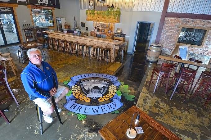 A man in a blue jacket sits on a stool in a brewery with the Antietam Brewery logo painted on the floor.