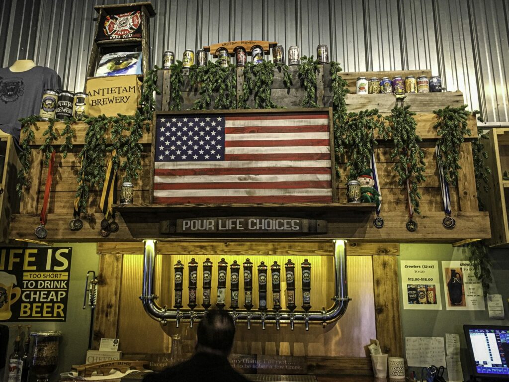 A brewery bar featuring a wall-mounted American flag, rows of beer taps, and shelves decorated with beer cans, garlands, and signs, with a person seated in the foreground.