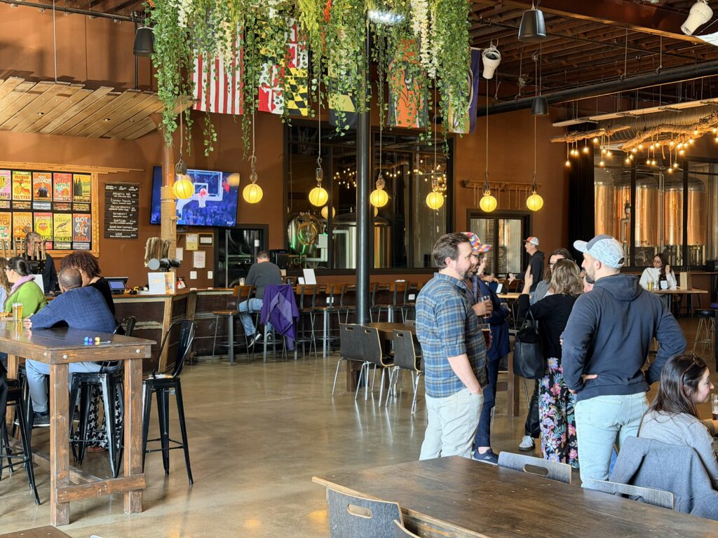 Patrons socialising in a brewery with wooden tables, bar seating, and hanging greenery, featuring a wall menu and a television showing a basketball game.