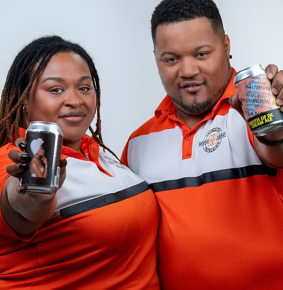 A man and a woman wearing matching orange, white, and black shirts hold up cans of beer.
