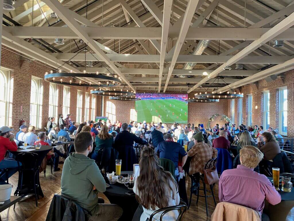 A crowd of people sitting at tables in a large hall with exposed beams, watching a soccer match on a large screen at the front of the room.