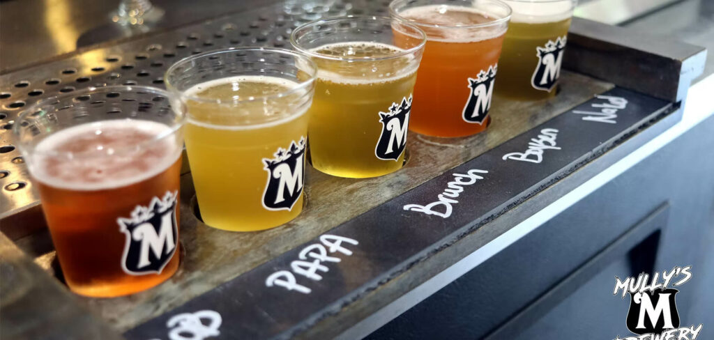 A flight of four craft beers in plastic cups, labeled on a black board with “Para,” “Brunch,” and “Boysen.”