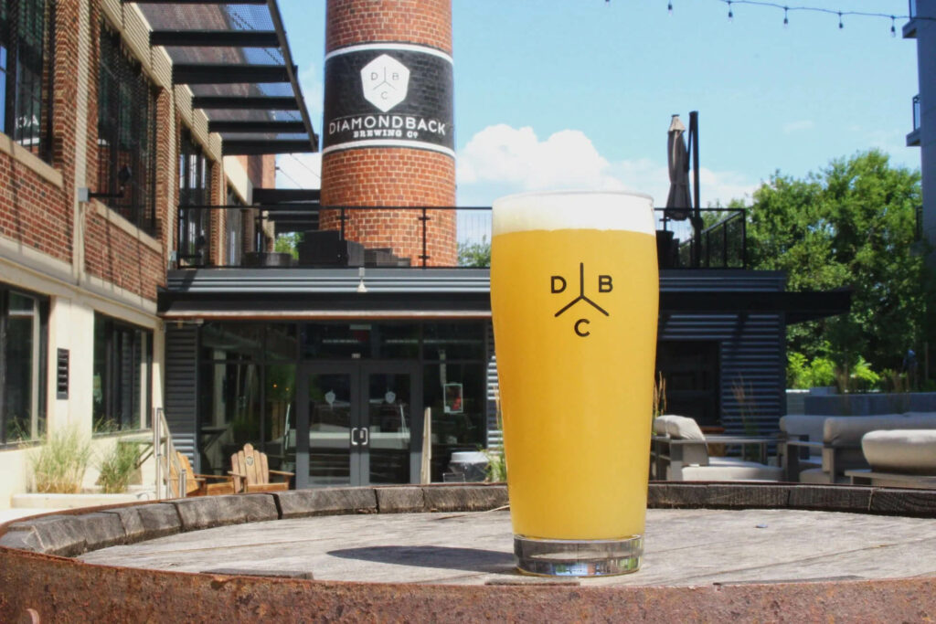 A glass of hazy beer with the Diamondback Brewing Co. logo sits on a wooden barrel in front of the brewery.