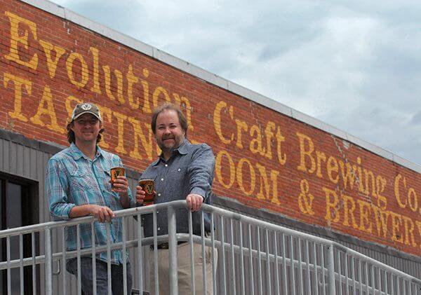 Two men holding glasses of beer stand on a metal balcony in front of a brick building with “Evolution Craft Brewing Co.” painted on the wall.