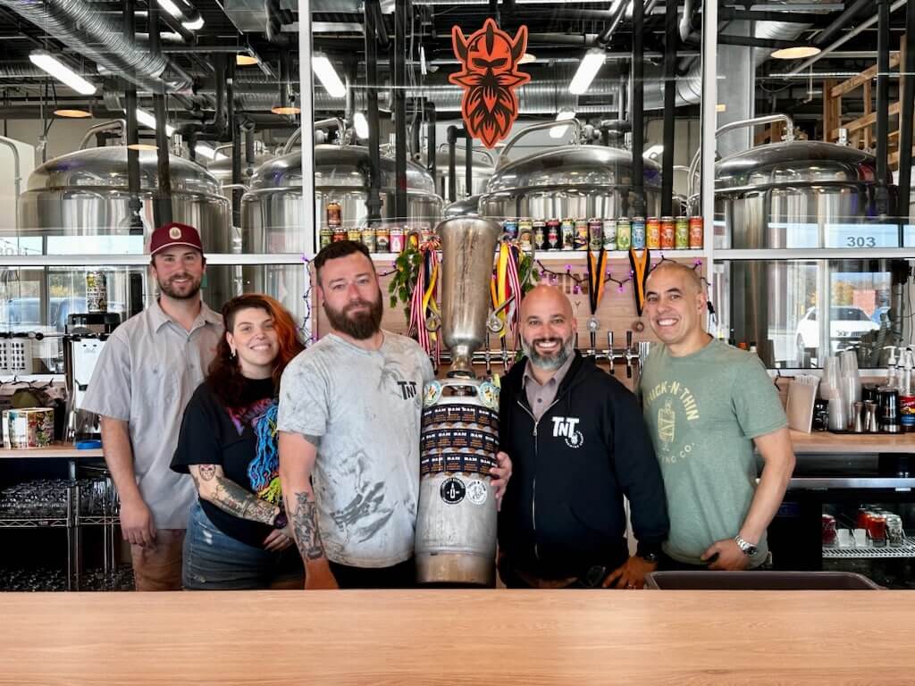 Five people standing behind a brewery bar, with the person in the center holding a tall trophy made from a beer keg.