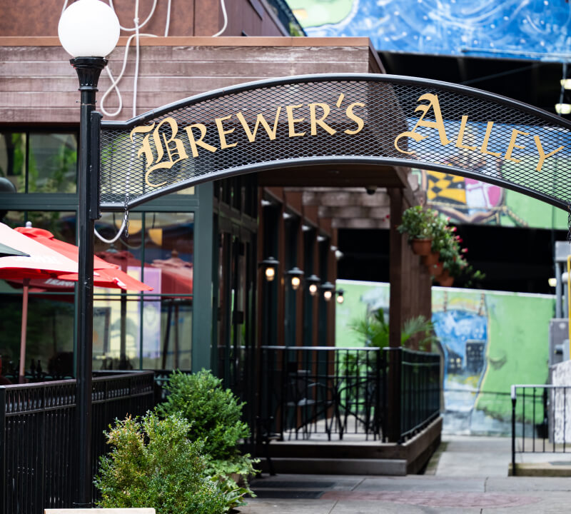 An arched metal sign reading “Brewer’s Alley” hanging above the entrance to a restaurant patio.