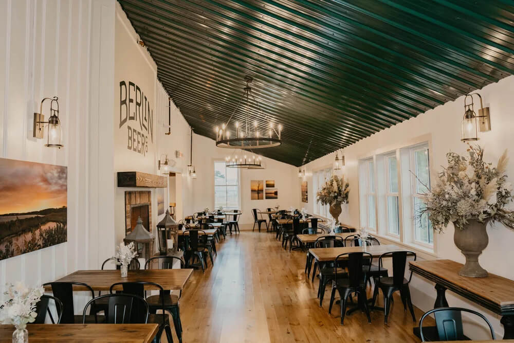 Interior of a restaurant with wooden tables, black chairs, and a long green sloped ceiling featuring the text “BERLIN BEER” on the wall.