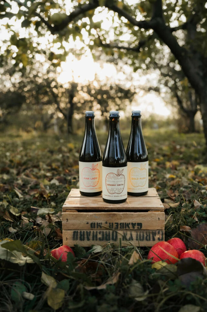 Three bottles of cider labeled Pink Lady, Granny Smith, and Gold Rush standing on a wooden crate in an orchard.