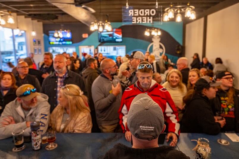 A group of people socializing at a brewery bar, with a man in a grey baseball cap seen from behind in the foreground.