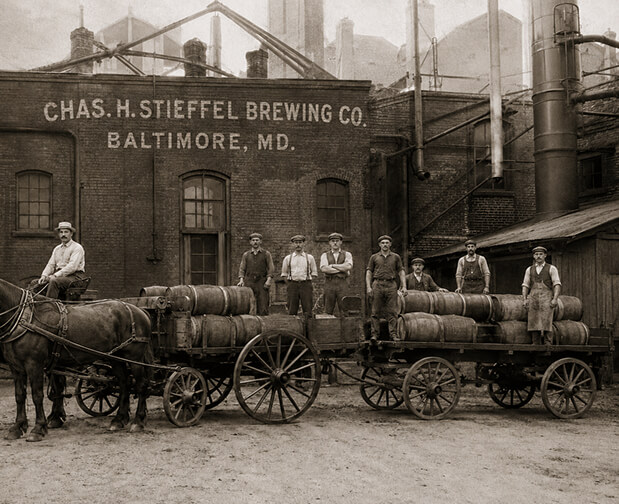 Seven men stand beside two horse-drawn wagons loaded with wooden barrels in front of the Chas. H. Stieffel Brewing Co. in Baltimore.