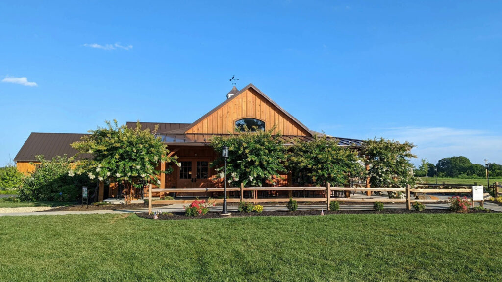 A large wooden barn-style building with a covered patio, surrounded by a low wooden fence and green lawn under a clear blue sky.