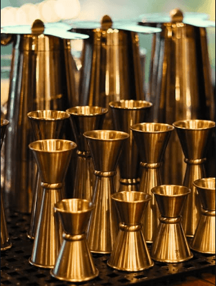 A collection of gold-toned metal cocktail jiggers arranged on a dark service mat.