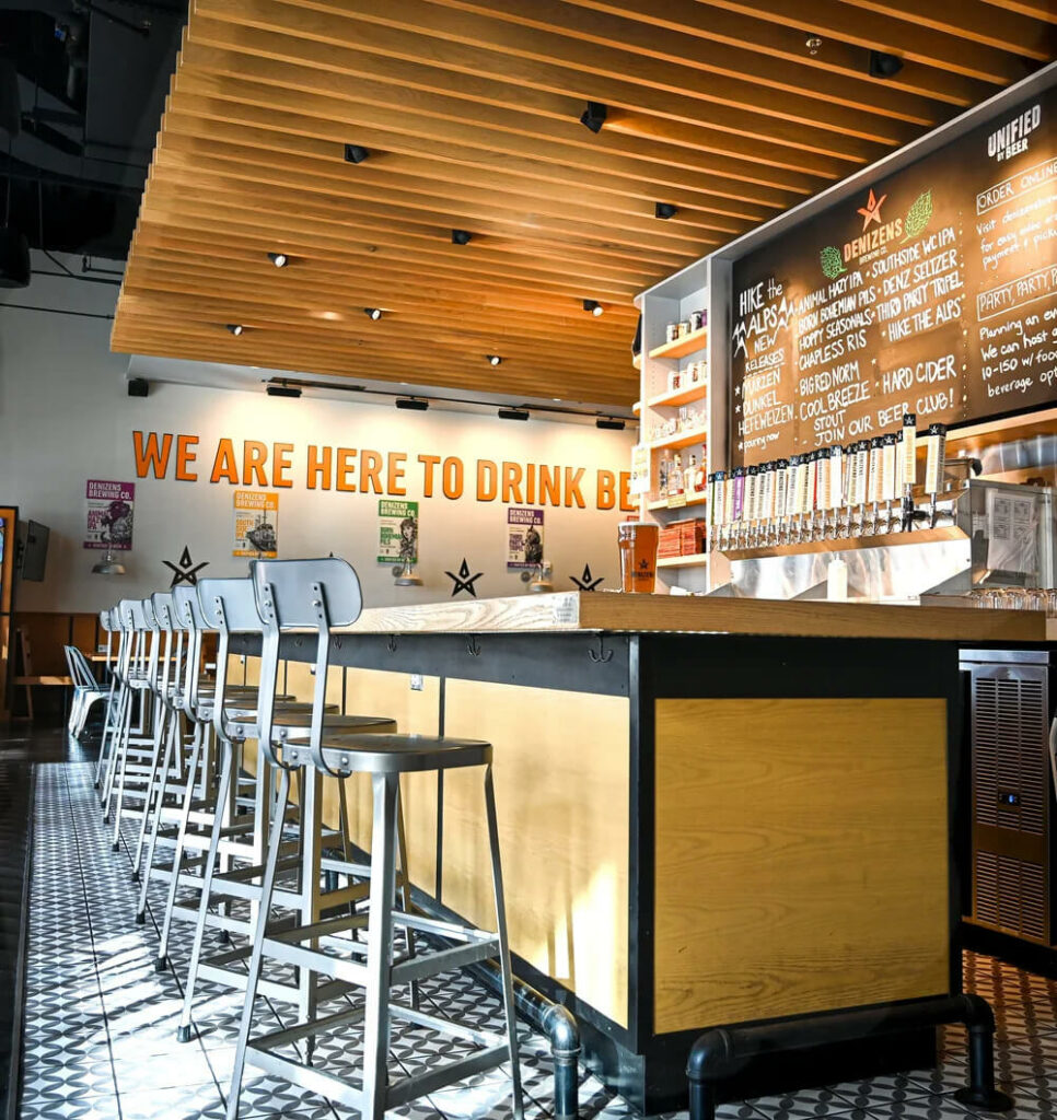 Modern brewery bar with metal stools, a wooden counter, and a large chalkboard menu listing beers behind the taps, with the sign “WE ARE HERE TO DRINK BEER” on the wall.