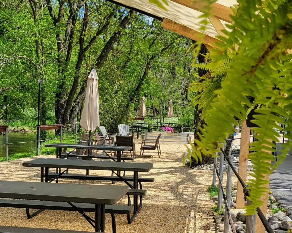 Outdoor patio seating with picnic tables and chairs along a tree-lined creek.