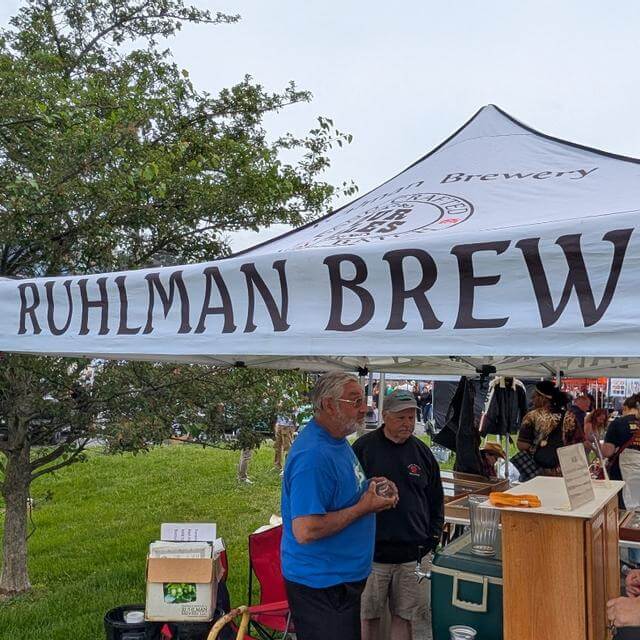 Two men stand under a Ruhlman Brew tent at an outdoor market.