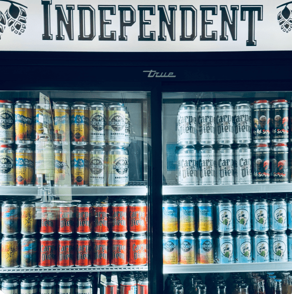 A glass-front refrigerator filled with shelves of craft beer cans, featuring an “INDEPENDENT” sign at the top.
