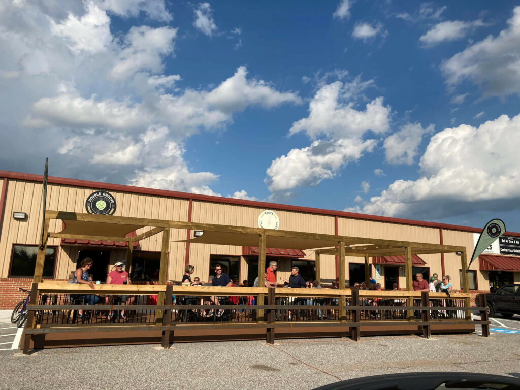 People sitting at tables on an outdoor wooden patio attached to a tan industrial building under a blue sky with scattered clouds.