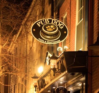 An oval sign for Pub Dog Pizza & Drafthouse hangs above the entrance to a brick building at night.