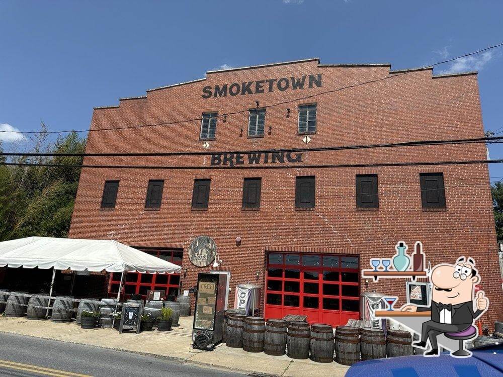 The brick building of Smoketown Brewing with a white tent, a row of barrels, and an illustrated mascot of a bartender in the foreground.