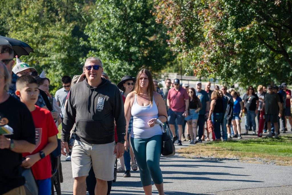 A crowd of people walk along a paved path in a park on a sunny day.