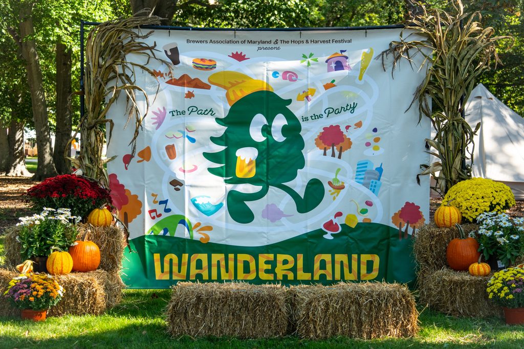 Festival banner reading “Brewers Association of Maryland & The Hops & Harvest Festival present The Path is the Party WANDERLAND” featuring a cartoon pine tree character holding a beer, surrounded by hay bales and potted mums.