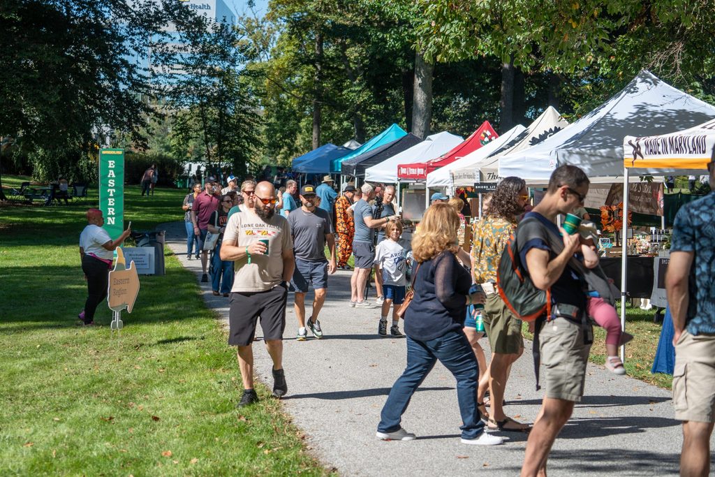 People stroll along a path lined with vendor tents at an outdoor market in a park.