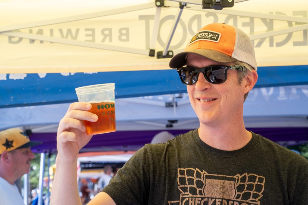 A man wearing a baseball cap and sunglasses holds up a plastic cup of beer at an outdoor event.